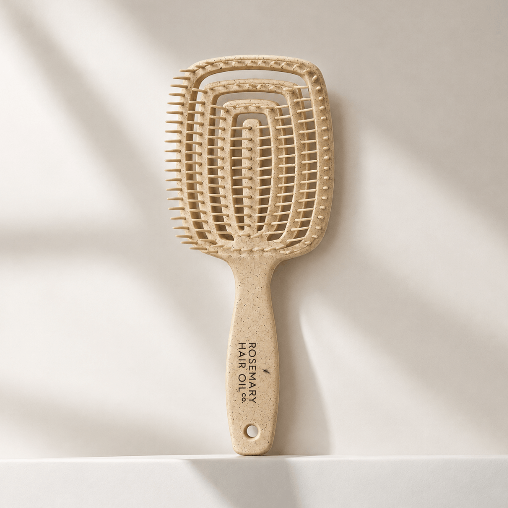 A natural hairbrush with a rectangular head and a long handle, standing upright on a white surface against a light beige background with soft diagonal shadows. The handle is printed with 'ROSEMARY HAIR OIL CO. Beige hairbrush with 'Rosemary Hair Oil Co.' branding on a beige background, detangle brush, tangle teaser, flexi brush